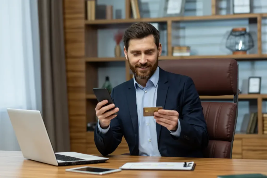 Geschäftsmann sitzt im Büro am Schreibtisch und benutzt Handy und Kreditkarte.