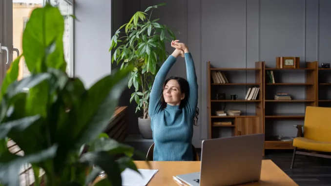 Eine Frau sitzt am Schreibtisch vor ihrem Laptop und streckt sich.