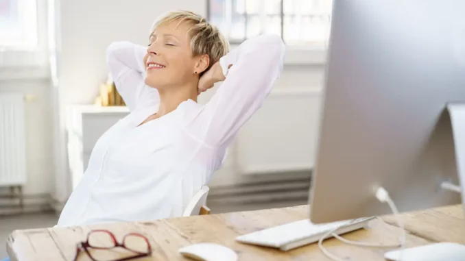Frau sitzt leicht zurückgelehnt vor ihrem Schreibtisch und hat dabei die Augen geschlossen. Sie macht eine Pause, um danach wieder effektiv zu arbeiten.