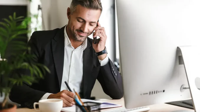 Ein Mann im Business-Outfit sitzt im Büro am Schreibtisch und führt ein geschäftliches Telefonat.