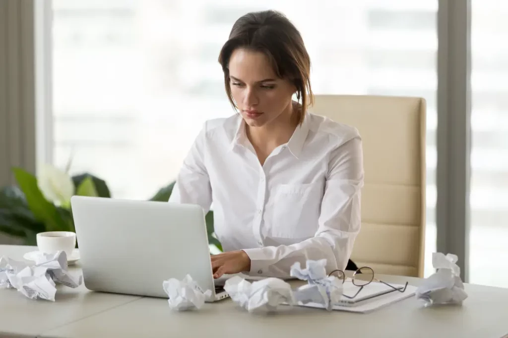 Frau im Business-Outfit sitzt am Schreibtisch und versucht zu am Laptop zu arbeiten. Um den Laptop herum liegen zerknitterte, weiße Blätter.