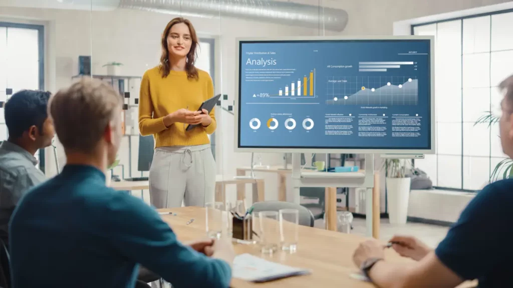 Eine Frau zeigt im Büro während eines Verkaufsgespräches ein digitales interaktives Whiteboard mit Wachstumsanalysen, Diagrammen, Statistiken und Daten.