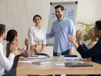 Ein Mann begrüßt während einer Teamversammlung im Büro eine neue Mitarbeiterin. Das Team klatscht zur Begrüßung.