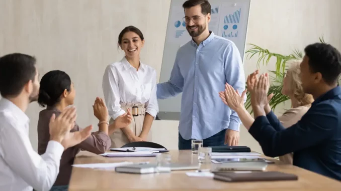 Ein Mann begrüßt während einer Teamversammlung im Büro eine neue Mitarbeiterin. Das Team klatscht zur Begrüßung.