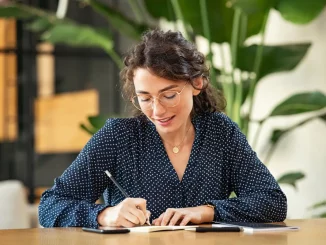 Eine Frau sitzt im Büro am Schreibtisch und schreibt in einem Notizbuch.