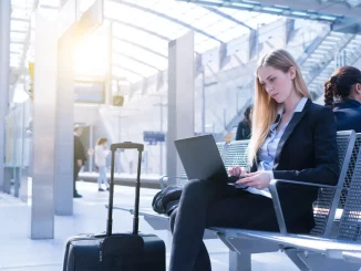 Eine Frau im Business-Outfit sitzt wartend am Bahnhof und arbeitet am Laptop.