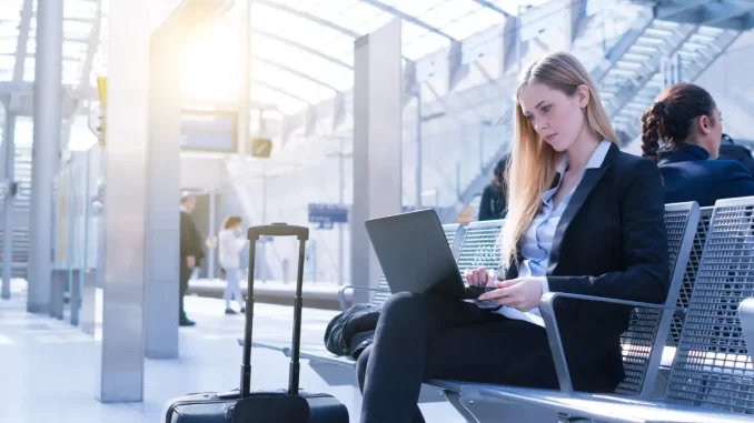 Eine Frau im Business-Outfit sitzt wartend am Bahnhof und arbeitet am Laptop.