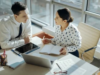 Ein Mann und eine Frau im Business-Outfit sitzen im Büro am Schreibtisch und planen den Tag gemeinsam in einem Planer.