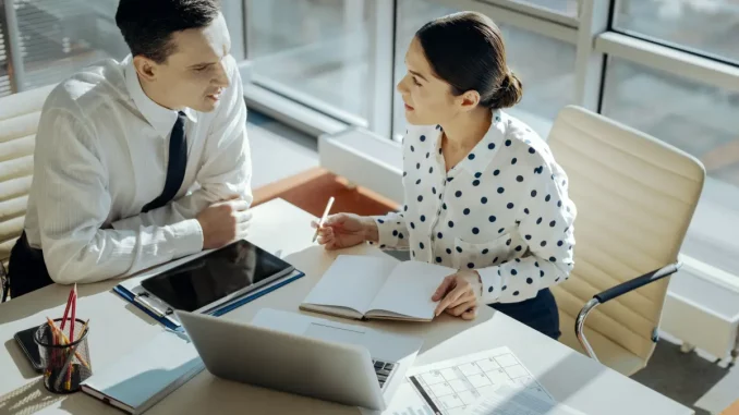 Ein Mann und eine Frau im Business-Outfit sitzen im Büro am Schreibtisch und planen den Tag gemeinsam in einem Planer.