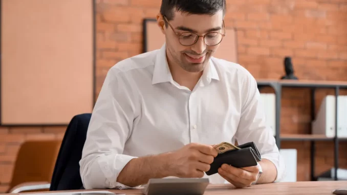 Ein Mann im Business-Outfit sitzt im Büro an seinem Schreibtisch und nimmt Geld aus seiner Geldbörse.