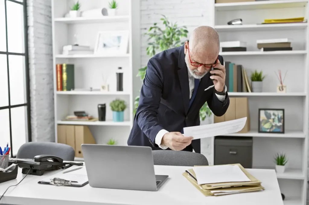 Ein Mann mittleren Alters im Business-Outfit mit Brille prüft einen Kontoauszug, während er in einem modernen Büro telefoniert.