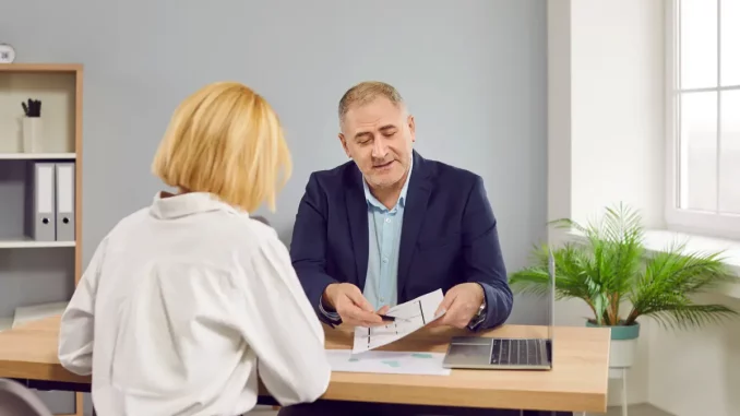 Auf dem Bild sitzen ein Mann und ein Frau an einem Schreibtisch. Der Mann zeigt der Frau einen Grundriss von einem Büro.