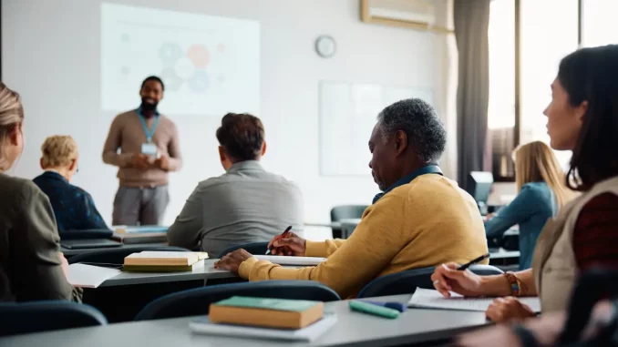 Ein Schulungsraum mit mehreren Schreibtischen an den unterschiedlich alte Personen sitzen, mit Blick auf eine digitale Tafel. Vor der Tafel steht ein Dozent.
