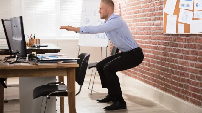 Ein Mann im Business-Outfit macht Kniebeuge vor dem Schreibtisch.
