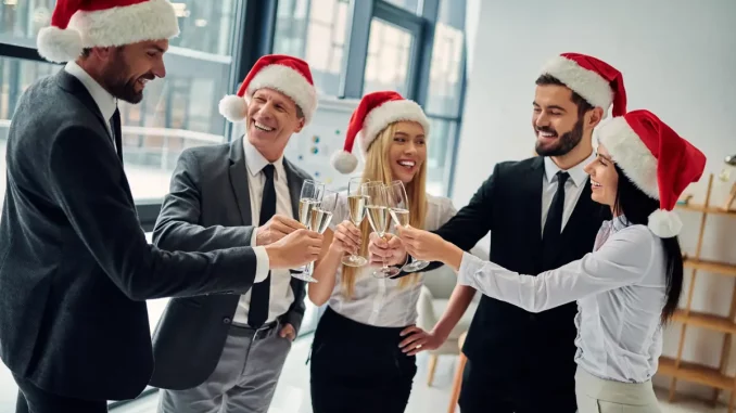 Fünf Kollegen im Business-Outfit mit Weihnachtsmann-Mützen stehen im Halbkreis und prosten sich mit einem Glas Sekt zu.