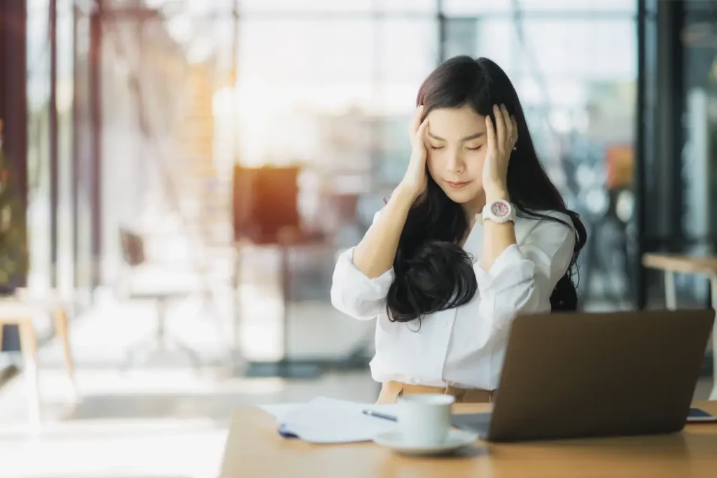 Frau mit langen Haaren sitzt im Business-Outfit am Schreibtisch. Sie wirkt konzentriert, hält sich die Hände an die Schläfen, Augen sind geschlossen.