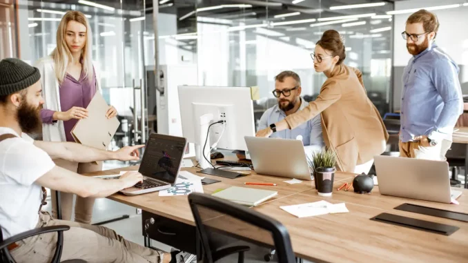 Zwei Männer sitzen am Schreibtisch, während sie von drei weiteren Kollegen unterbrochen werden, die sich neben Ihnen an den Schreibtisch gestellt haben.