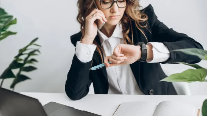 Nahmaufnahme einer Frau im Business-Outfit, die am Schreibtisch sitzt. Sie telefoniert und schaut auf ihre Uhr am Handgelenk. Sie wirkt abgelenkt von ihrer eigentlichen Arbeit.