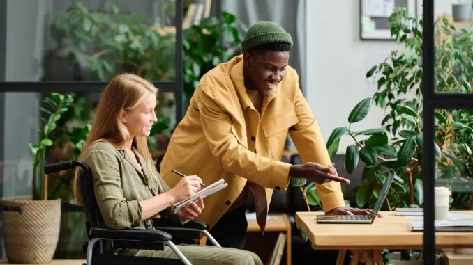 Bild von einer Frau im Rollstuhl vor ihrem Schreibtisch. Neben ihr steht ein Kollege, der auf ihren Arbeitsbildschirm zeigt.