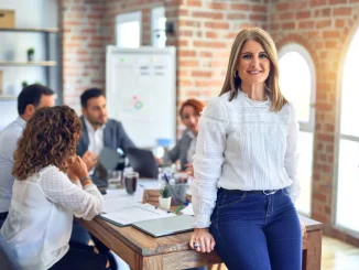 Eine Gruppe von vier Kollegen sitzt gemeinsam an einem Tisch im Büro. Vorn am Tisch angelehnt, steht eine Frau mit einem Lächeln im Gesicht.