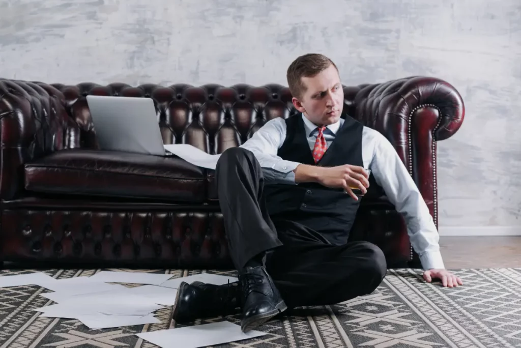 Ein Mann im Business-Outfit sitzt auf dem Boden vor einer Couch, mit einem Whiskeyglas in der Hand. Um ihn herum liegen lose Dokumente.
