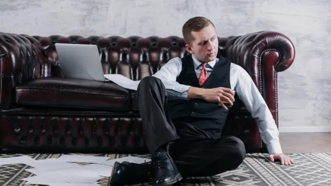 Ein Mann im Business-Outfit sitzt auf dem Boden vor einer Couch, mit einem Whiskeyglas in der Hand. Um ihn herum liegen lose Dokumente.