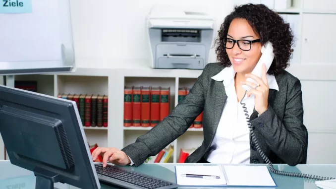 Eine Frau sitzt am Schreibtisch, den Telefonhörer in der linken Hand und die rechte Hand auf der Computertastatur. Sie scheint eine Telefonkonferenz zu organisieren.