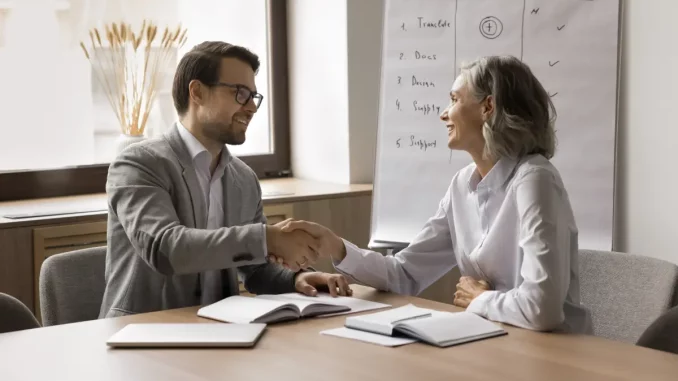 Ein Mann und eine Frau mittleren Alters sitzen im Büro am Schreibtisch und reichen sich nach dem Bewerbungsgespräch die Hände.