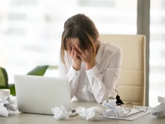 Eine Frau sitzt vor ihrem Latop am Schreibtisch und hält die Hände vor ihr Gesicht. Um den Laptop liegen zerknüllte Blätter.