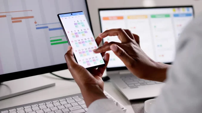 Auf dem Bild ist zu sehen, wie eine Frau ein Handy festhält. Auf dem Display ist der geöffnete Kalender zu sehen, mit farblich unterschiedlich markierten Terminen.
