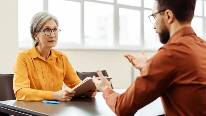 Eine Frau und ein Mann im Hemd sitzen sich an einem Tisch gegenüber. Die Frau hört dem Mann aufmerksam zu, während der Mann leicht gestikuliert, um seinen Worten wahrscheinlich Ausdruck zu verleihen.