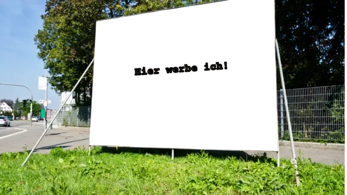 Ein weißes Werbeschild mit der Aufschrift "Hier werbe ich!" steht auf einer Grünfläche neben einer Vekehrsstraße.