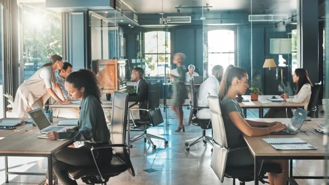 Blick in ein hektisches Großraumbüro mit zahlreichen Arbeitnehmern an ihren Schreibtischen, umgeben von Computerbildschirmen und Büroausstattung. Die Szene vermittelt eine stressige Arbeitsumgebung.