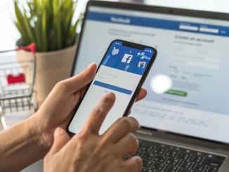 Bild zum Thema 'Lukrative E-Mail-Listen über Facebook aufbauen': Ein Laptop mit geöffneter Facebook-Seite, davor eine Hand mit einem Smartphone, auf dem ebenfalls Facebook zu sehen ist, und ein kleiner Einkaufswagen neben dem Laptop.