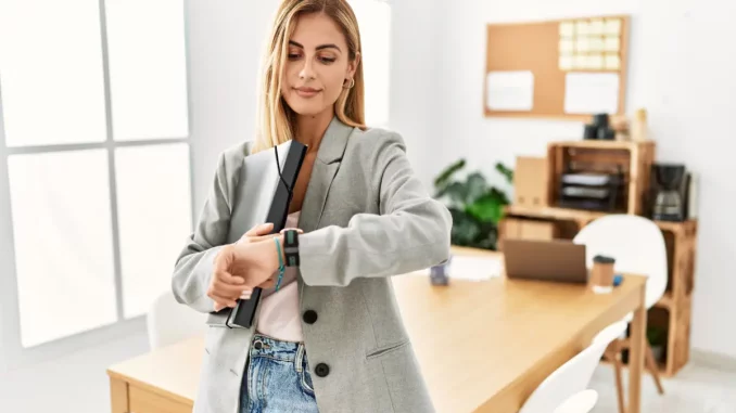 Frau im Jacket steht im Büro und schaut auf die Uhr.