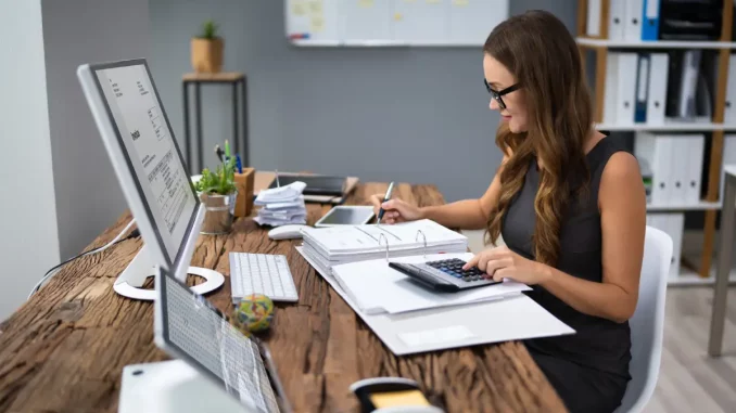 Frau im Business-Outfit sitzt am Schreibtisch im Büro und rechnet die Mahngebühren einer unbezahlten Rechnung aus.