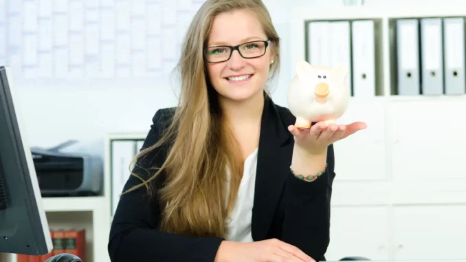 Nahaufnahme einer Frau im Business-Outfit am Schreibtisch im Büro mit Sparschwein in der Hand.