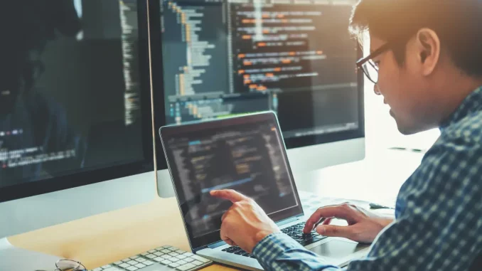 Ein Mann sitzt konzentriert an einem Schreibtisch vor einem Computerbildschirm, auf dem leicht verschwommene Codes zu sehen sind. Dieses Bild illustriert einen Artikel über IT-Karrieren, indem es den Fokus auf die tägliche Arbeit eines Programmierers legt.