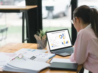 Eine Frau sitzt konzentriert am Schreibtisch, vor ihr ein Laptop mit einem Leistungsdiagramm, umgeben von Firmenunterlagen und einem Taschenrechner, symbolisch für strategische Unternehmensplanung zur Vermeidung von Insolvenz.
