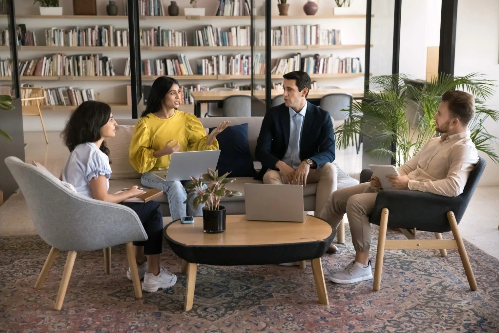Besprechung mit Laptops in einer Büroecke mit Sofa, Sessel, Teppich und Bücherregalen.